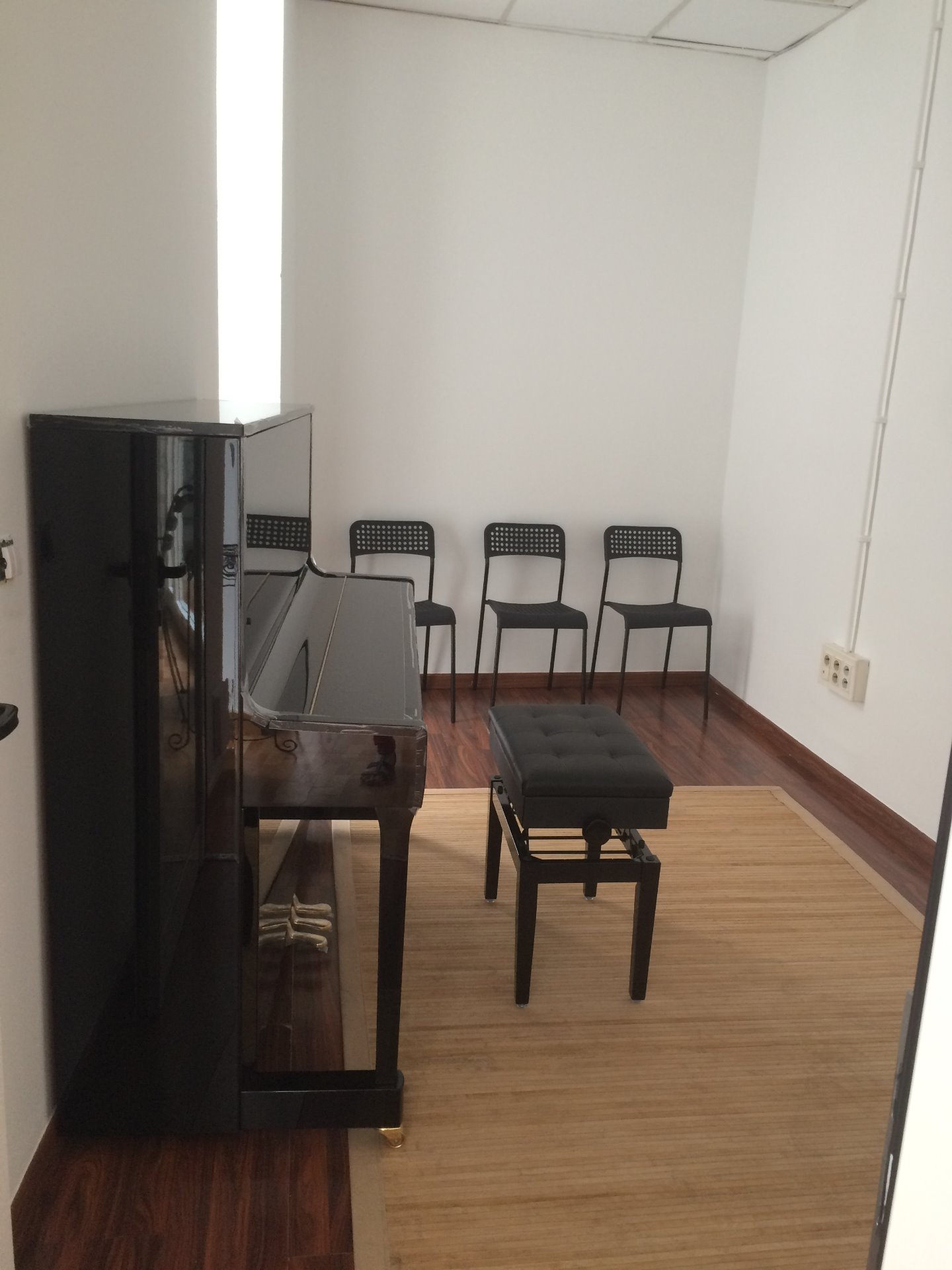 Pequeña sala de práctica con un piano vertical, tres sillas negras, un banco de piano y suelo de madera